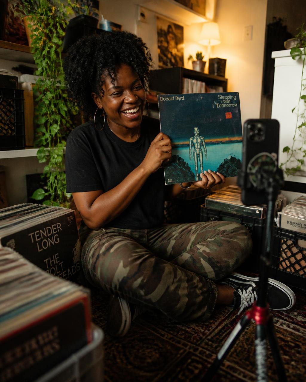A record collector laughing while holding up a rare album sleeve to a phone on a tripod in her apartment